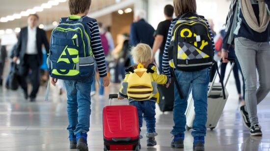 Three caucasian boys walk through an airport terminal. The youngest boy by many years is pulling a small red suitcase. The two older boys are wearing backpacks and holding his hand.