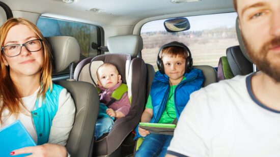 A caucasian family is in their car. The mother is wearing glasses and is holding a book. The focus is on a young boy in the back seat, middle. To his right is a young baby in a forward-facing carseat.