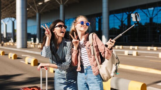 Two girls smile while taking a selfie using a selfie-stick outside of an airport temrinal. They're wearing sunglasses and blue jeans.