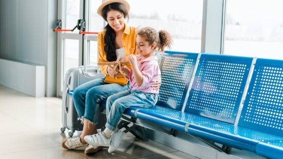 A mother sits on a bench at an airport with her daughter. They are playing with a wooden model airplane and smiling.