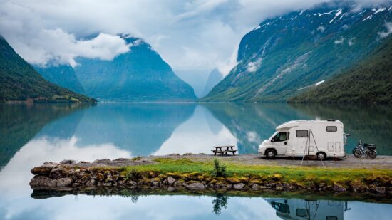 A perfectly calm fjord with an RV (recreational vehicle) and mopeds on a small peninsula in the foreground. The outdoor scene is very peaceful.