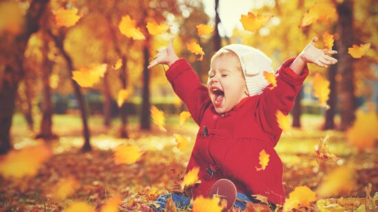 A thrilled young girl throws leaves in an autumn setting. She's wearing a hat and red jacket and has her mouth wide open from laughing